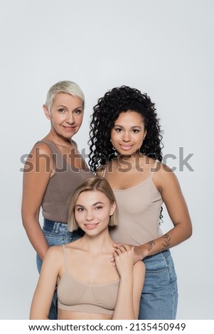 Smiling interracial women looking at camera near young blonde friend isolated on grey