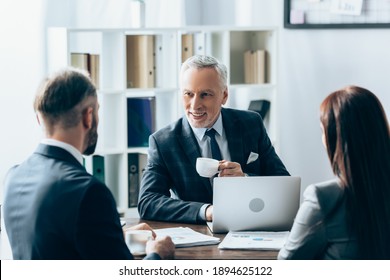 Smiling investor with cup of coffee looking at businesspeople on blurred foreground near papers and laptop - Shutterstock ID 1894625122