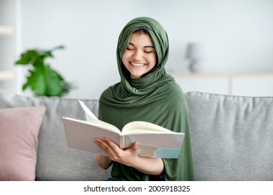 Smiling Islamic Teen Girl In Hijab Reading Book, Enjoying Interesting Story At Home. Cheerful Muslim Adolescent In Headscarf Looking Through Textbook, Getting Ready For Exam, Studying Remotely