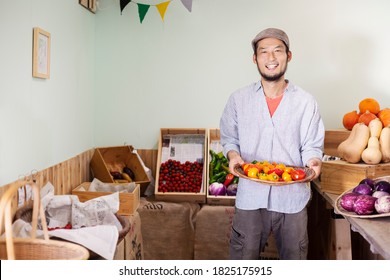 Smiling Japanese man wearing cap standing in farm shop, holding bowl with fresh peppers - Shutterstock ID 1825175915