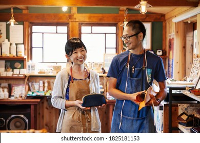 Smiling Japanese woman and man wearing blue apron and glasses standing in a leather shop. - Shutterstock ID 1825181153