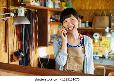 Smiling Japanese Woman Wearing Apron Standing In A Leather Shop, Talking On Her Mobile Phone.