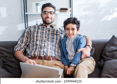 smiling jewish father with laptop and son looking at camera in apartment 