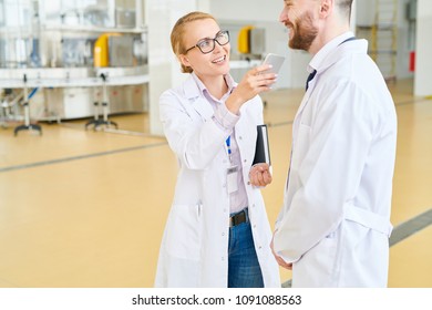 Smiling Journalist Wearing White Coat Using Dictaphone On Smartphone While Talking To Cheerful Bearded Owner Of Soy Milk Factory, Interior Of Spacious Production Department On Background