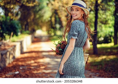 Smiling joyful happy caucasian redhead girl in straw hat and fluttering dress walks alone in autumn park on sunny warm day while holds straw handbag. Indian summer concept.