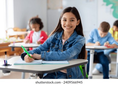 Smiling junior school girl sitting at desk in classroom, writing in notebook, posing and looking at camera. Group of diverse classmates studying on background