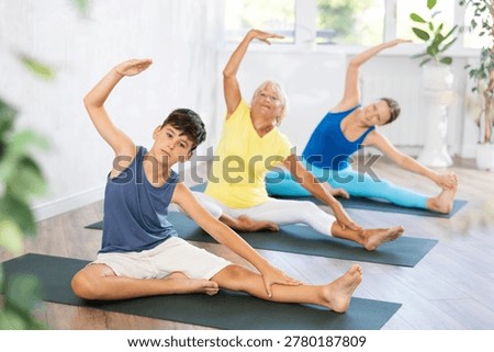 Smiling juvenile boy doing revolved head to knee pose of yoga in gym room with other persons