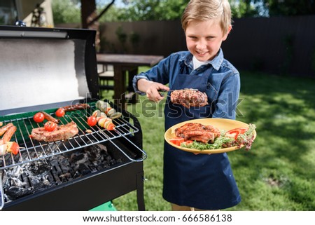 smiling kid boy in apron preparing tasty stakes on barbecue grill outdoors