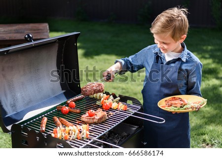 smiling kid boy in apron preparing tasty stakes on barbecue grill outdoors