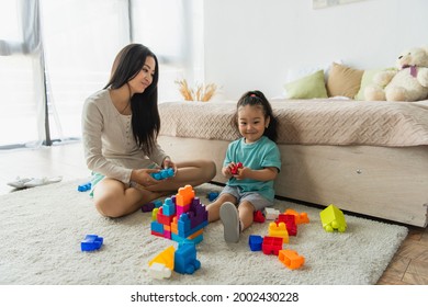 Smiling Kid Holding Building Block Near Asian Mother In Bedroom