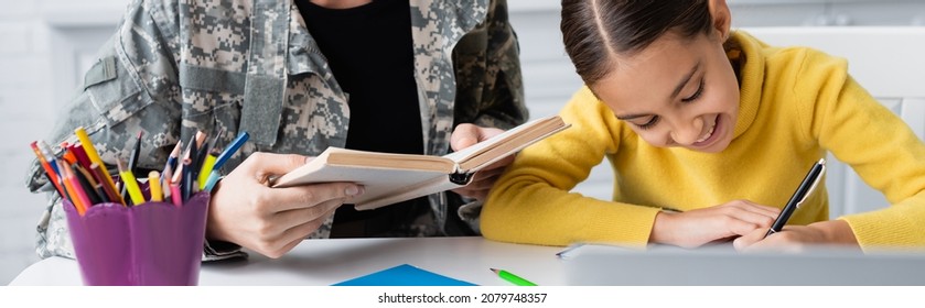 Smiling kid holding pen near mother in military uniform with book near laptop at home, banner - Powered by Shutterstock