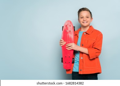 Smiling Kid Holding Penny Board And Looking At Camera On Blue Background