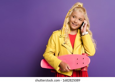 Smiling Kid Holding Penny Board And Talking On Smartphone On Purple Background