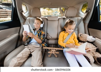 Smiling Kids With Smartphone And Book Sitting Near Toys On Back Seat Of Car 