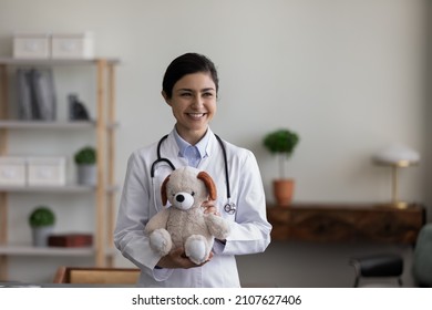 Smiling Kind Young Indian Female Pediatrician General Practitioner Doctor With Teddy Bear Toy In Hands, Looking In Distance Waiting For Small Patients For Checkup Meeting In Modern Clinic Office.