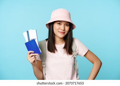 Smiling korean girl tourist, showing passport and tickets for flight, booking hotel, going on trip, voyage, standing over blue background