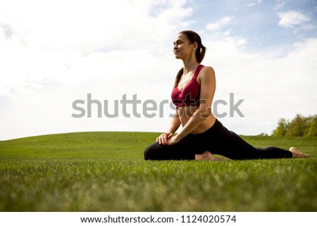 Smiling lady is doing stretching exercises on green lawn. She is sitting on one hip and straighten other while closing eyes with delight. Copy space in right side