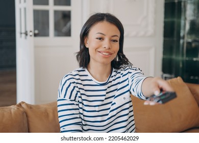 Smiling Lady Enjoying Movie In Living Room. Happy Spanish Woman Sitting On Leather Sofa. Young Woman Is Watching Television With Remote Controller. Rest And Relaxation, Weekend At Home.