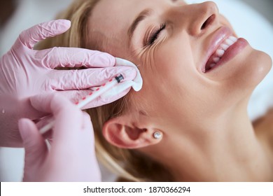 Smiling lady lying with her eyes closed during the rejuvenation injection administered by a professional dermatologist