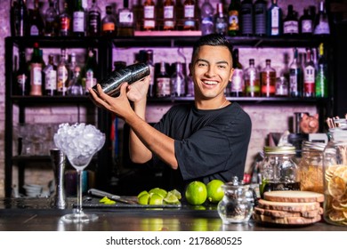 A smiling Latin American bartender mixes liquids with a cocktail shaker in front of a pub bar. - Powered by Shutterstock