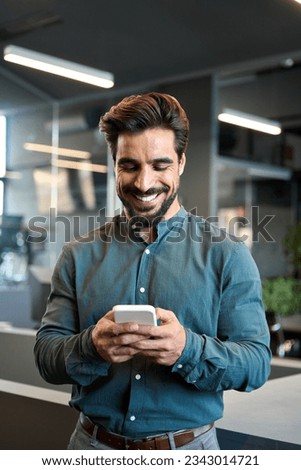 Smiling Latin business man entrepreneur using smartphone standing in office. Happy young businessman holding cellphone working on mobile phone making digital payment standing in office, vertical.