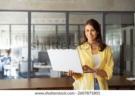 Smiling latin hispanic female marketing manager, professional it specialist working, browsing at laptop computer standing at workplace in office. Young woman employee using pc for business. Copy space