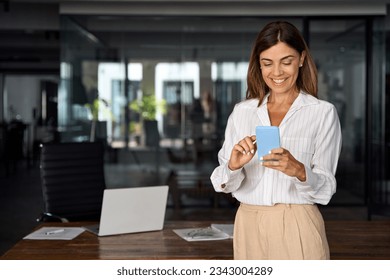 Smiling Latin Hispanic mature adult professional business woman using mobile phone cellphone. European businesswoman CEO holding smartphone using fintech application standing at workplace in office.