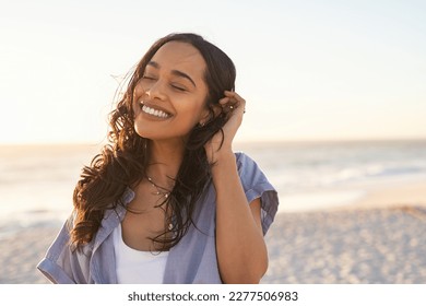 Smiling latin hispanic woman relaxing on beach with closed eyes at sunset. Beautiful mixed race woman enjoying wind fluttering hair. Charming young woman breathing fresh air at summer beach.
