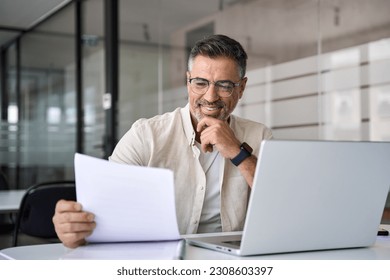 Smiling Latin or Indian male business man accountant analyst holding documents, work at laptop computer doing online trade market tech research. Focused Hispanic businessman with paperwork in office.