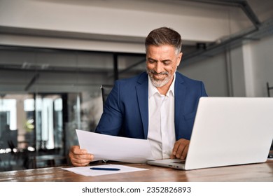 Smiling Latin or Indian male business man accountant analyst holding documents, work at laptop computer doing online trade market tech research. Focused Hispanic businessman with paperwork in office. - Powered by Shutterstock