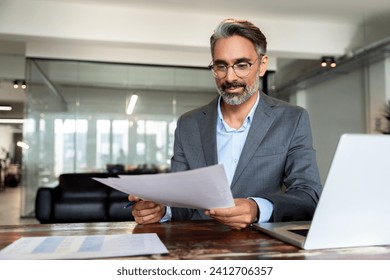 Smiling Latin or Indian male business man accountant analyst holding documents, work at laptop computer doing online trade market tech research. Focused Hispanic businessman with paperwork in office. - Powered by Shutterstock