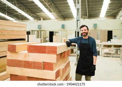 Smiling Latin Man Posing Next To A Pile Of Wooden Boards. Male Worker Doing His Job In A Warehouse And Woodshop