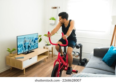 Smiling latin man riding his stationary bike at home while watching a workout video on the living room tv - Powered by Shutterstock