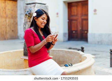 smiling latin woman with flower in her hair sitting by a fountain typing on a phone