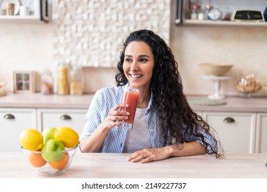 Smiling latin woman hold glass of organic tomato juice, sitting at table in light kitchen interior, enjoying healthy beverage. Proper nutrilon, breakfast, health care concept