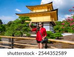 A smiling Latina tourist takes a photo with her phone in front of Kinkaku-ji, the Golden Pavilion, in Kyoto, Japan. She enjoys the scenic view while dressed casually in a red blouse and sunglasses.