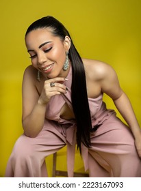 Smiling Latina Woman Model In Pink Jumper And Yellow Backdrop Sitting Down With Long Dark Hair 