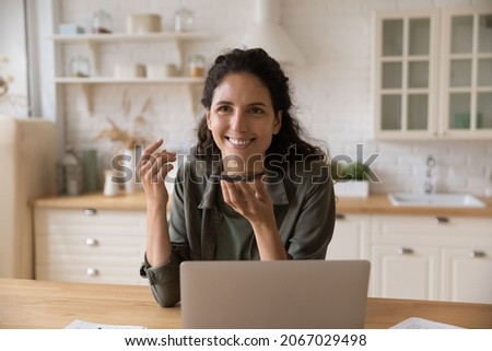 Smiling Latina woman sit at table with laptop holds smartphone talks on speakerphone, leave voicemail message chatting remotely enjoy personal or formal distant conversation. Modern ai tech concept