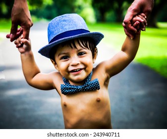 Smiling Latino Baby Wearing A Blue Hat And Bow Tie Walking Outside And Holding Hands With Adults