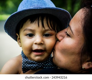 Smiling Latino Baby Wearing A Blue Hat And Bow Tie Being Kissed On Cheek By Female Adult Family Member