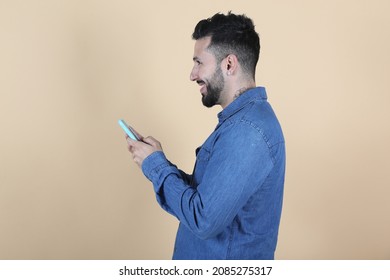 Smiling Latino Hispanic Man Stands In Profile While Holding His Phone With His Hands On Yellow Background