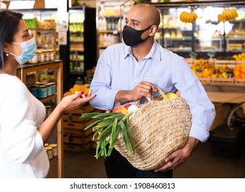 Smiling Latino Wearing Protective Mask To Prevent Viral Infection Standing With Purchases In Grocery Store, Friendly Talking To Woman..