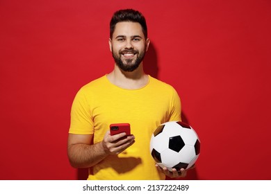 Smiling Laughing Beautiful Young Bearded Man Football Fan In Yellow T-shirt Cheer Up Support Favorite Team Hold Soccer Ball Use Mobile Cell Phone Isolated On Plain Dark Red Background Studio Portrait