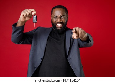 Smiling laughing young african american business man 20s wearing classic jacket suit standing pointing index finger on camera hold car keys isolated on bright red color background studio portrait