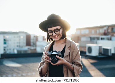 Smiling And Laughing Young Woman, Professional Photographer Stands On Top Of Rooftop In City On Warm Summer Evening, Makes Photos With Retro Vintage Analog Camera