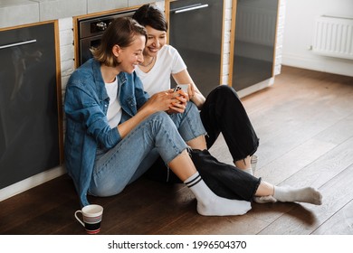 A smiling lesbian couple looking at the phone while sitting on the floor in the light kitchen - Powered by Shutterstock