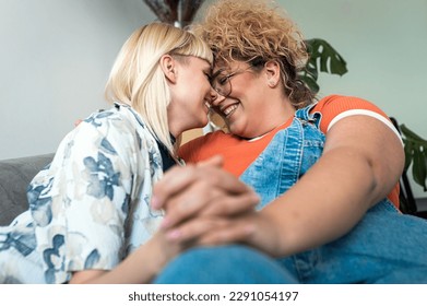 A smiling lesbian couple sits on the sofa, hugging each other, leaning their foreheads together and holding hands at home. - Powered by Shutterstock