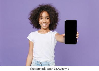 Smiling little african american kid girl 12-13 years old in white t-shirt isolated on violet wall background studio portrait. Childhood lifestyle concept. Hold mobile phone with blank empty screen