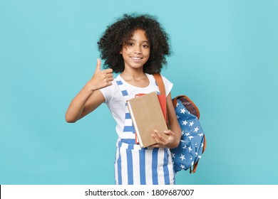 Smiling little african american kid schoolgirl 12-13 years old in striped clothes with backpack hold books, showing thumb up isolated on blue background studio. Childhood education in school concept.