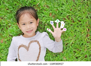 Smiling Little Asian Child Girl Lying On Green Grass Lawn With Showing White Stickers With Number One To Five On Her Fingers.